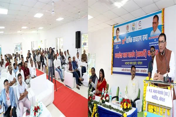 सभा कक्ष में बैठे श्रोता और फूलों से सजे मंच पर बोलते वक्ता; पीछे 28वीं वार्षिक साधारण सभा का बैनर