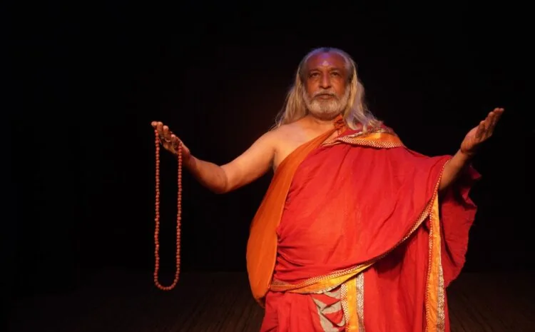 काले मंच पर नारंगी वस्त्र पहने अभिनेता माला लेकर संवाद करते हुए